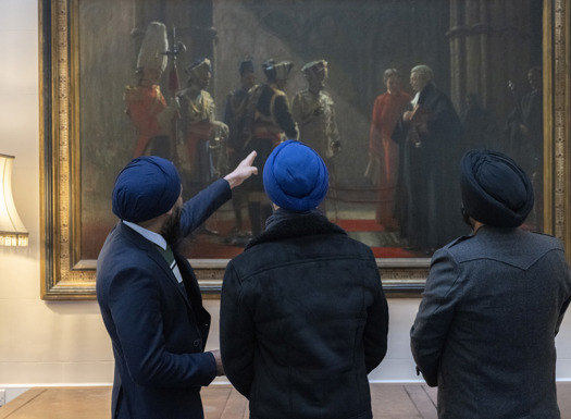 Three Sikh serving personnel with their backs to the camera, wearing blue turbans, pointing at a painting called Homage to the Fallen