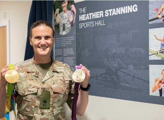 Heather Stanning holding her Olympic medals in front of her wall plaque inside the sports hall.