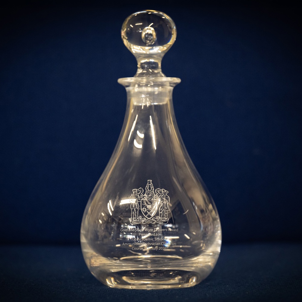 Clear glass decanter with a round stopper and an intricate emblem etched on its front against a dark blue background.