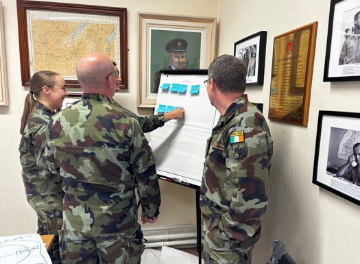 Four military personnel in camouflage placing sticky notes onto a flip chart. They are standing in front of various paintings and plaques