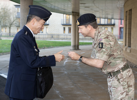 Major General Rowell, dressed in military working dress bows respectfully to his Japanese counterpart, whilst presenting him with a business card, as both men smile.