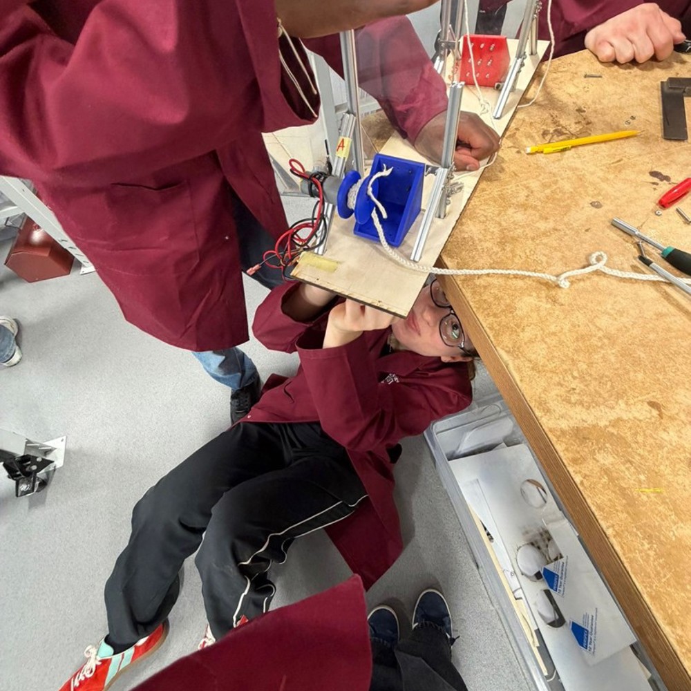 Student lying on the floor assembling an electronic project under a wooden table while another holds the device above.
