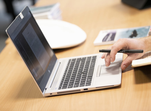 A laptop on a desk with a person using one hand to use the tracker pad. In their hand they hold a pencil