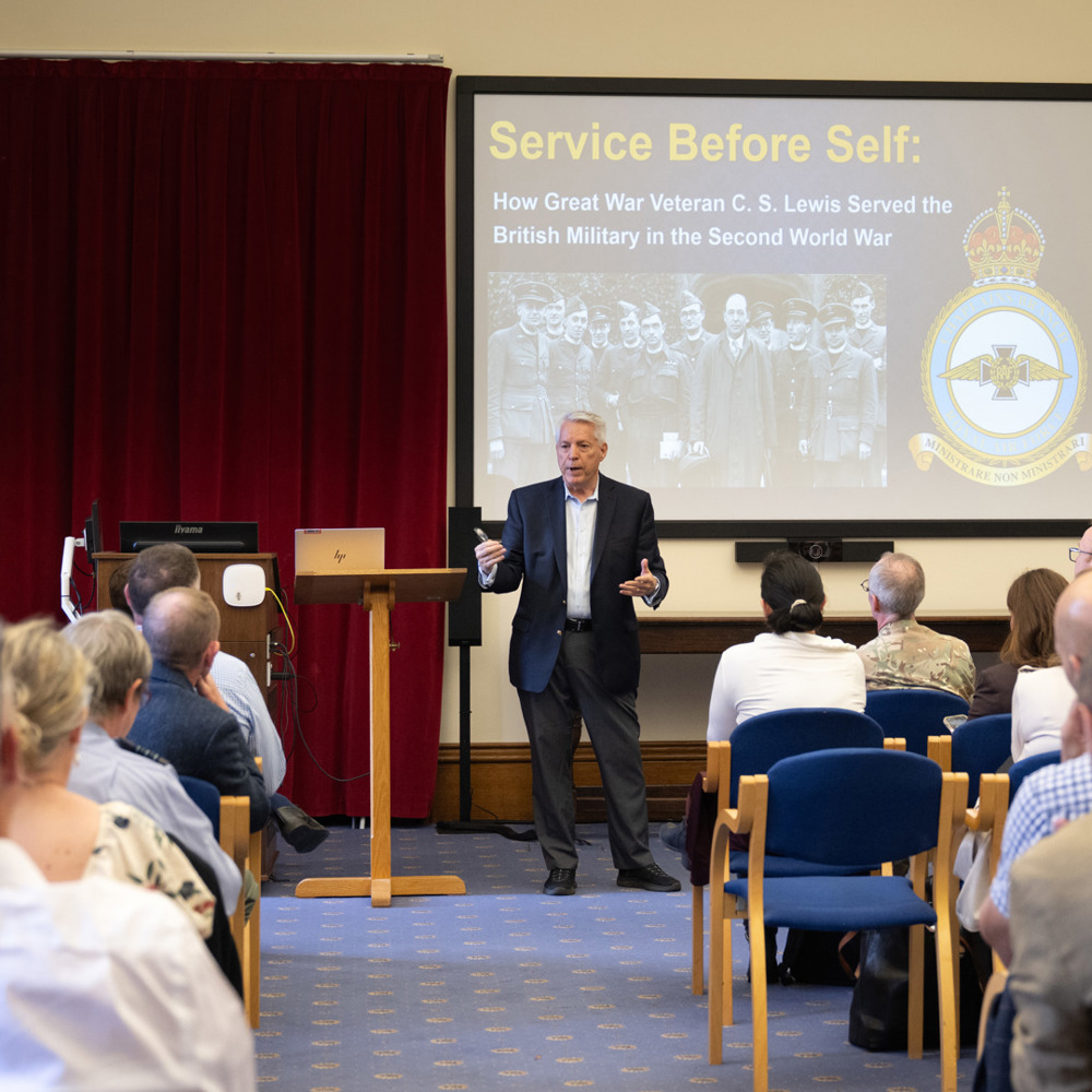 Speaker presenting a lecture titled 'Service Before Self' about C.S. Lewis's role in the British military during WWII to an attentive audience.
