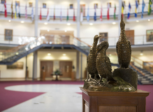 A gold statue of 3 cormorant birds inside a forum filled with multiple handing flags from around the world.