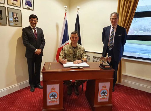 Army officer sat at a desk signing KIASS contract bookended by two individuals in suit and tie. 