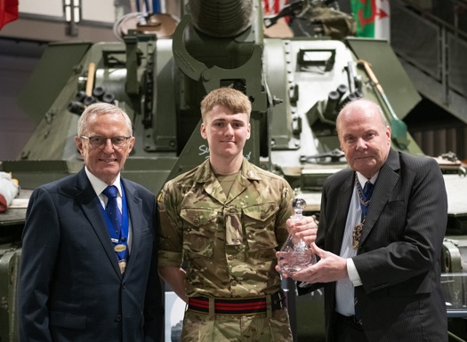 Three individuals stand in front of a military tank. The central figure wears a camouflage military uniform, while the two on either side are in formal attire with medals. The person on the right holds a glass trophy