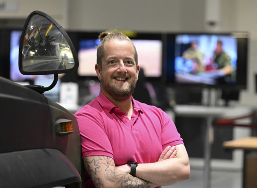 Civilian in pink shirt lent against an armoured vehicle with digital screens in the background.