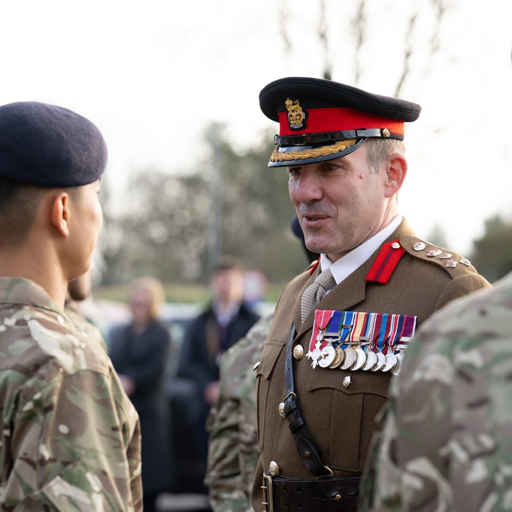A senior military officer in dress uniform with medals speaks to two soldiers in camouflage during an outdoor ceremony.