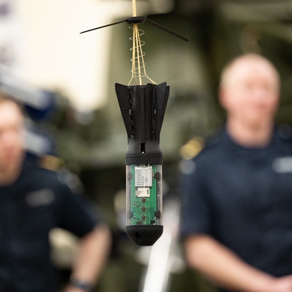Close-up of a small black sensor with visible circuit board and antenna, suspended in front of two blurred uniformed personnel.