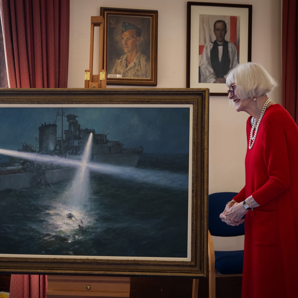 A woman in a red dress stands beside a large framed painting displayed on an easel. The painting shows a naval ship at night shining a bright searchlight onto the water, where a figure is visible in the illuminated waves. The scene takes place in a room with red curtains, wooden furniture, and framed artwork on the walls
