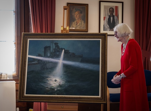 A woman in a red dress stands beside a large framed painting displayed on an easel. The painting shows a naval ship at night shining a bright searchlight onto the water, where a figure is visible in the illuminated waves. The scene takes place in a room with red curtains, wooden furniture, and framed artwork on the walls