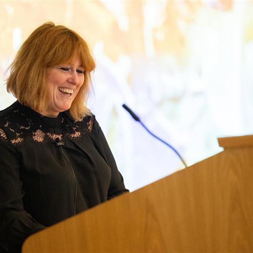 Woman in a black lace top speaking at a wooden podium with a microphone in a softly lit room.