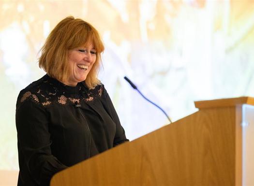 Woman in a black lace top speaking at a wooden podium with a microphone in a softly lit room.