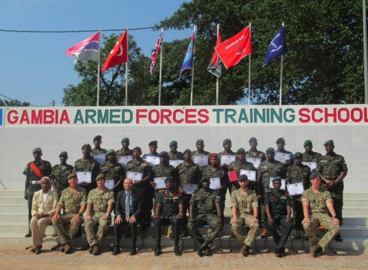 Group photo of Gambia Armed Forces trainees and instructors holding certificates in front of the training school sign and flags.