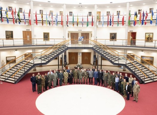 A group of personnel stood in a semi-circle surrounded by overhead international flags. 