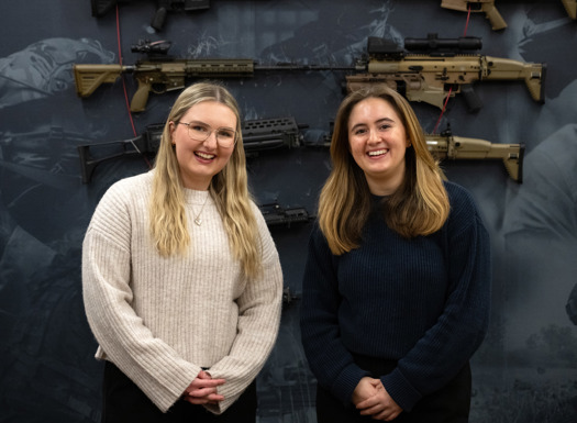 Two young women stood in front of a blue wall with guns, smiling