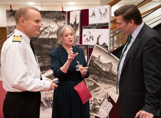 A navy officer, civilian and Minister for the Armed Forces talking with artwork in the background.