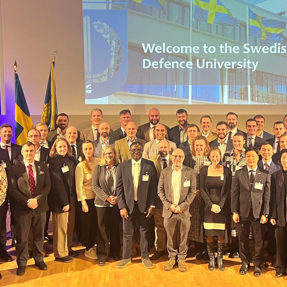 Group photo of diverse professionals standing on stage at the Swedish Defence University event with flags and a welcome message in the background.