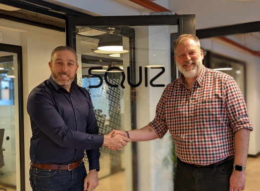 Two men in civilian dress shaking hands in front of a glass door with the company name 'Squiz'.
