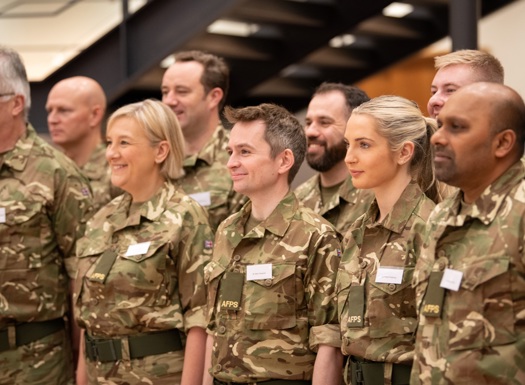  A group of smiling individuals in matching camouflage uniforms pose for a photo, all looking side on to the camera.