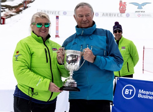 Cdr Nicky Cullen being presented with the Oliver Delaney Trophy in front of snowy ski slopes.