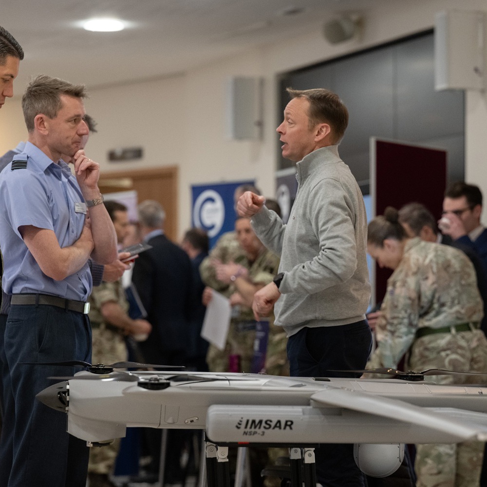 Military personnel and a civilian discussing near a drone displayed at an indoor exhibition or event.