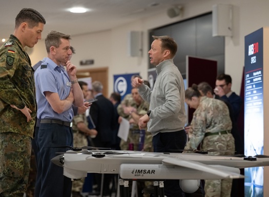 Military personnel and a civilian discussing near a drone displayed at an indoor exhibition or event.