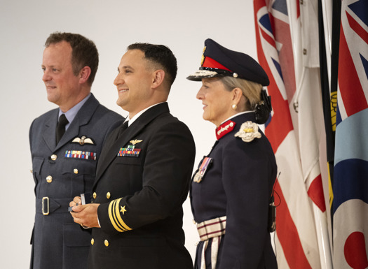 Three people in uniform with military flags in the background.