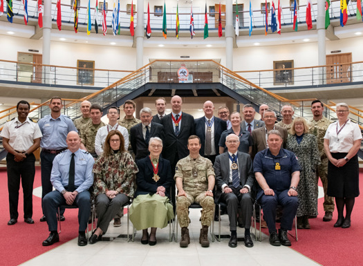 A mix of military British officers and civilians in a large grand hall with international flags hung in the background.